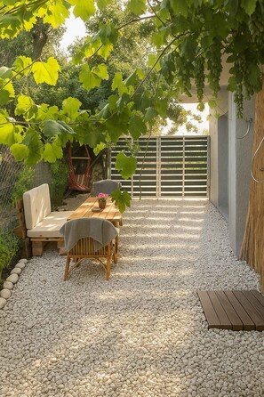 Un patio con mesa y sillas de madera, rodeado de grava, bajo un enrejado cubierto de hiedra.