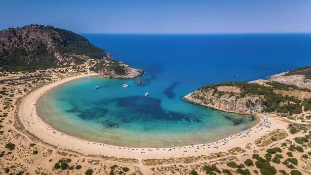 An aerial view of a coastal area with a sandy beach, clear blue waters, and rocky terrain.