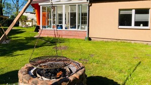 Un jardin avec un barbecue, une balançoire et une maison avec un toit rouge et de grandes fenêtres.