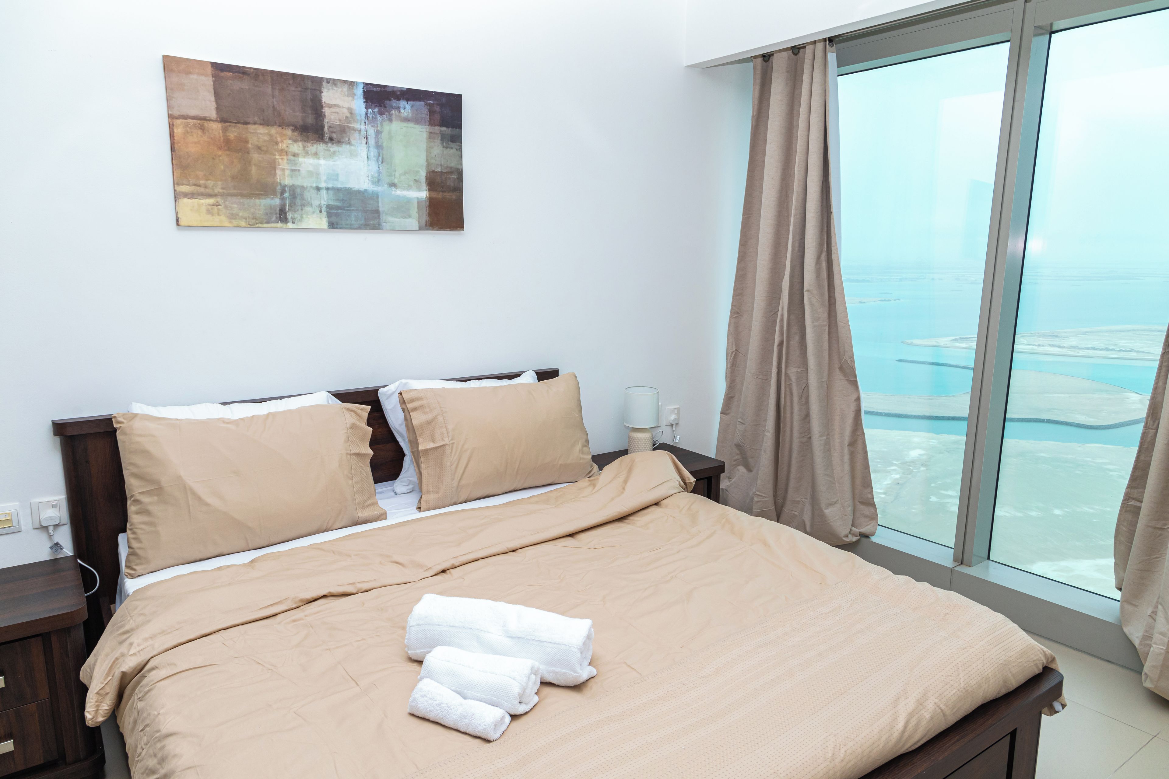 Habitación de hotel con una cama grande, una mesita de noche, una lámpara y vistas al mar a través de un ventanal amplio.