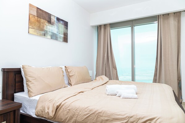 A hotel room with a large bed, a wooden nightstand, a painting on the wall, and a view of the sea through the window.