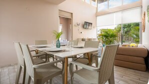 A dining area with a table set for four, surrounded by wicker chairs, a television mounted on the wall, and a large window offering a view of greenery outside. - Spondylus Bungalow (Canoas)