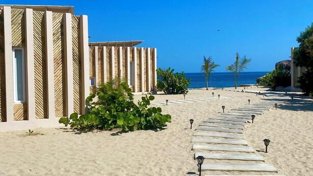 A beachside walkway leading to a building with vertical wooden slats, a clear view of the ocean, and a row of small lamps along the path. - Spondylus Bungalow (Canoas)