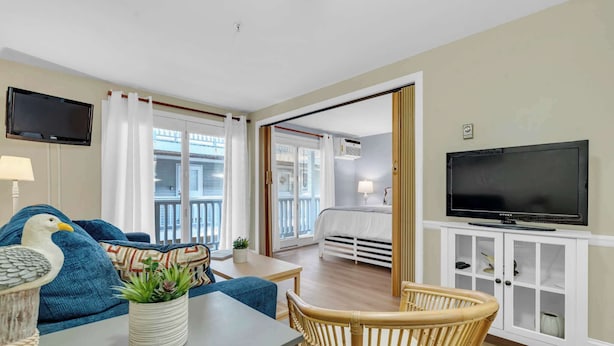 Une chambre d’hôtel moderne dotée d’un balcon, d’une télévision, d’un coin repas avec une table et des chaises, et d’un lit avec une tête de lit blanche.