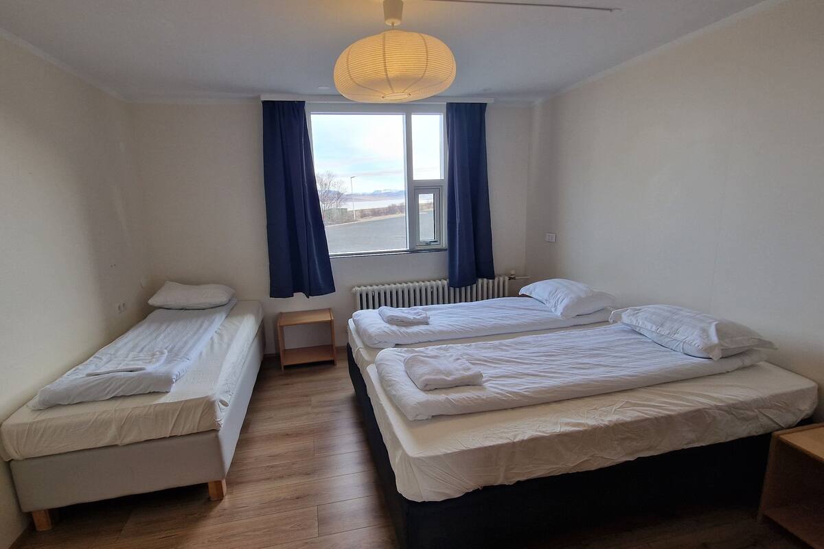 A room with two single beds, a wooden floor, a radiator, a window with curtains, and a hanging light fixture.