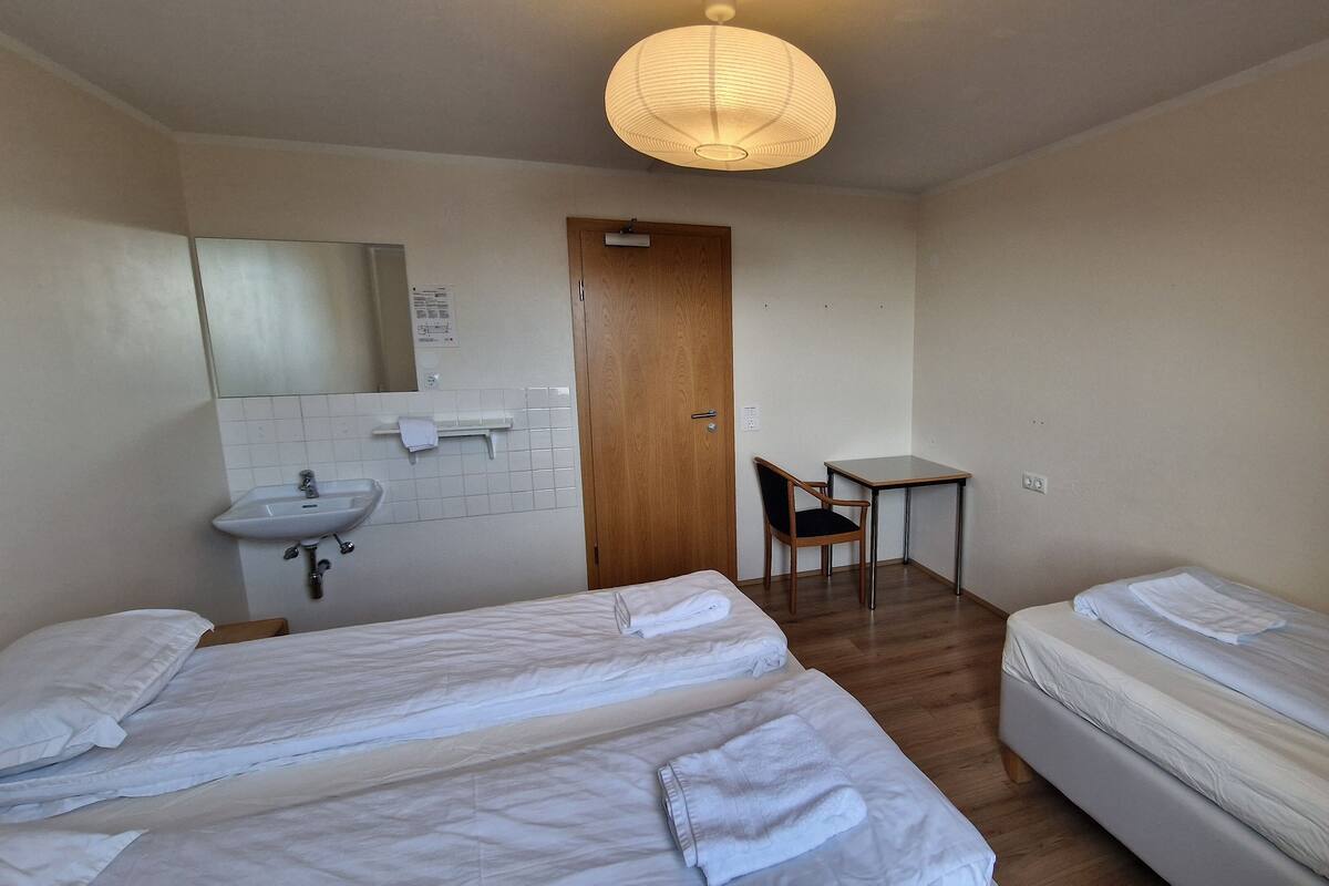 A small, clean room with two beds, a desk, a chair, and a sink.