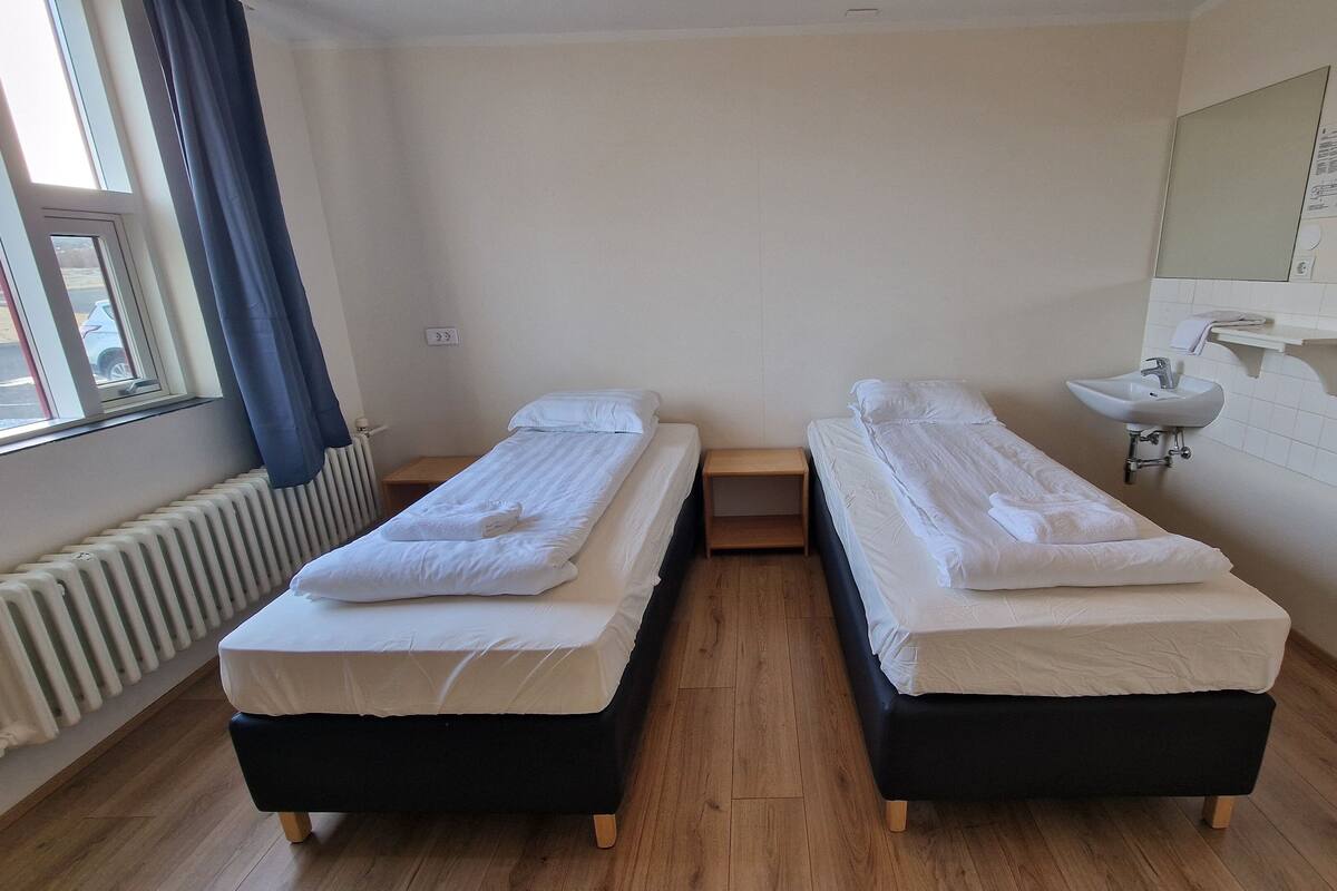 A small, clean room with two single beds, a wooden nightstand, a white sink, and a window with curtains.