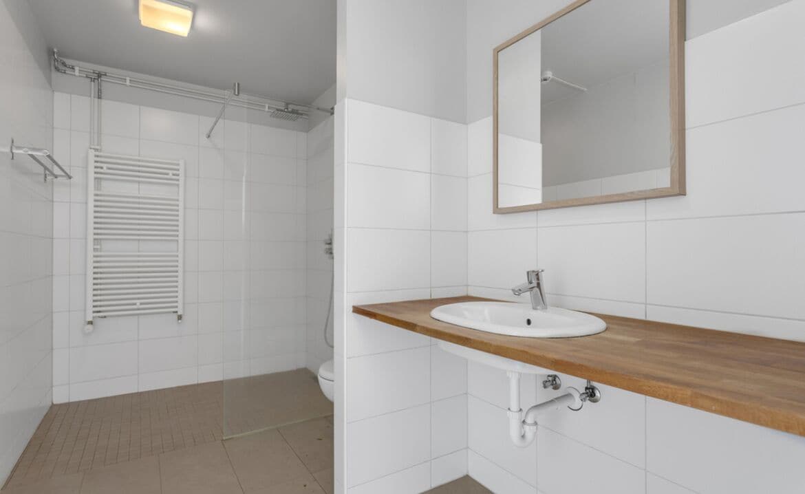 A modern bathroom with white tiles, a wooden vanity, a mirror, and a shower.