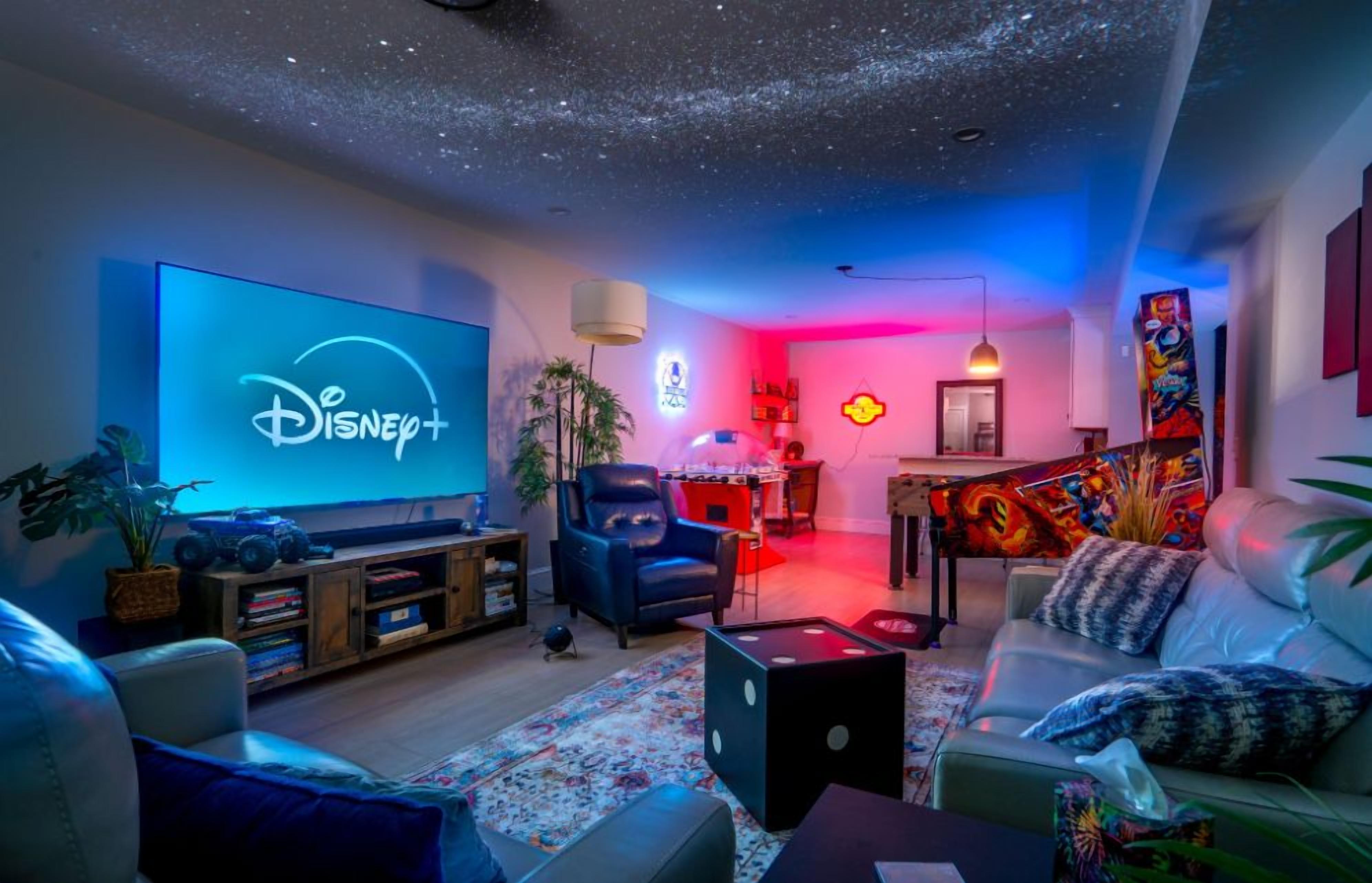 A living room with a flat-screen TV displaying the Disney+ logo, a gaming chair, a sofa, a coffee table, and a bookshelf.