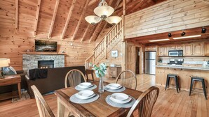 A wooden cabin interior with a dining table set for four, a fireplace, a kitchen with a microwave, and a staircase leading to an upper level.