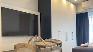 A modern hotel room with a large wardrobe, a TV mounted on the wall, a baby crib, and a bed. - Superior Suite | Zotos Luxury Living #2 (Thessaloniki)