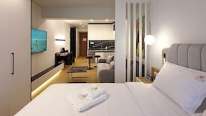 A modern hotel room with a neatly made bed, a desk with a laptop, a sofa, and a kitchen area in the background. - Superior Suite | Zotos Luxury Living #2 (Thessaloniki)
