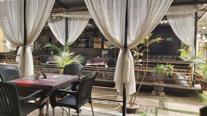 Un área de estar al aire libre con mesas y sillas, cubierta por cortinas blancas y rodeada de plantas en macetas.