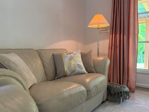 Un sofá de cuero color beige con cojines, una lámpara de pie y una ventana con cortinas.