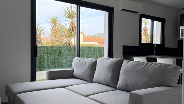 A modern living room with a grey sectional sofa, large windows, and a view of palm trees and buildings. - Comfortable gîte ideally located (Saint-Martin-d'Ardèche)