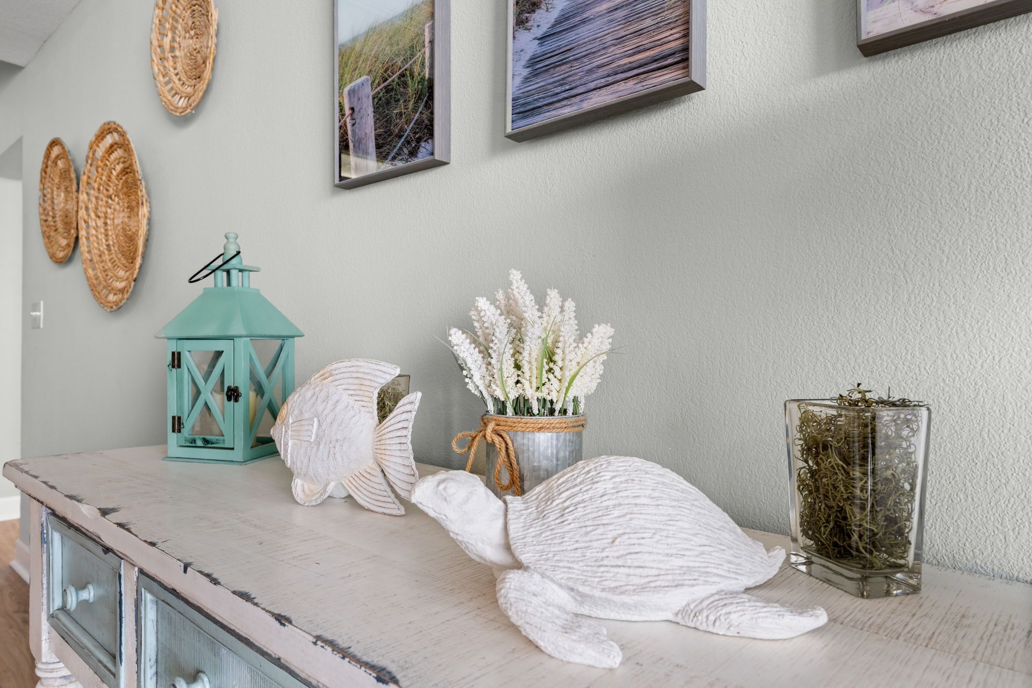 A room with a rustic wooden dresser, a white turtle sculpture, a teal lantern, a vase with white flowers, and a glass container with greenery.