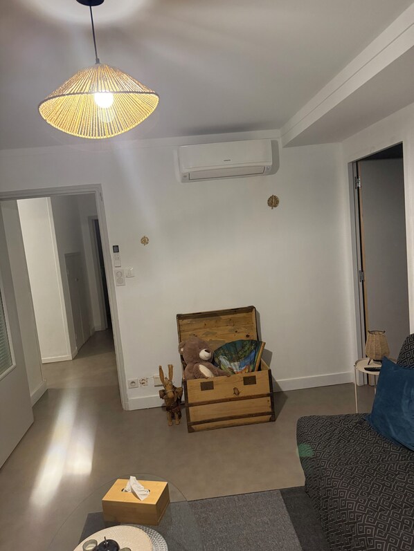 Una habitación con un cofre de madera que contiene un osito de peluche y un cuadro, un sofá con funda estampada, una lámpara colgante y un equipo de aire acondicionado en la pared.