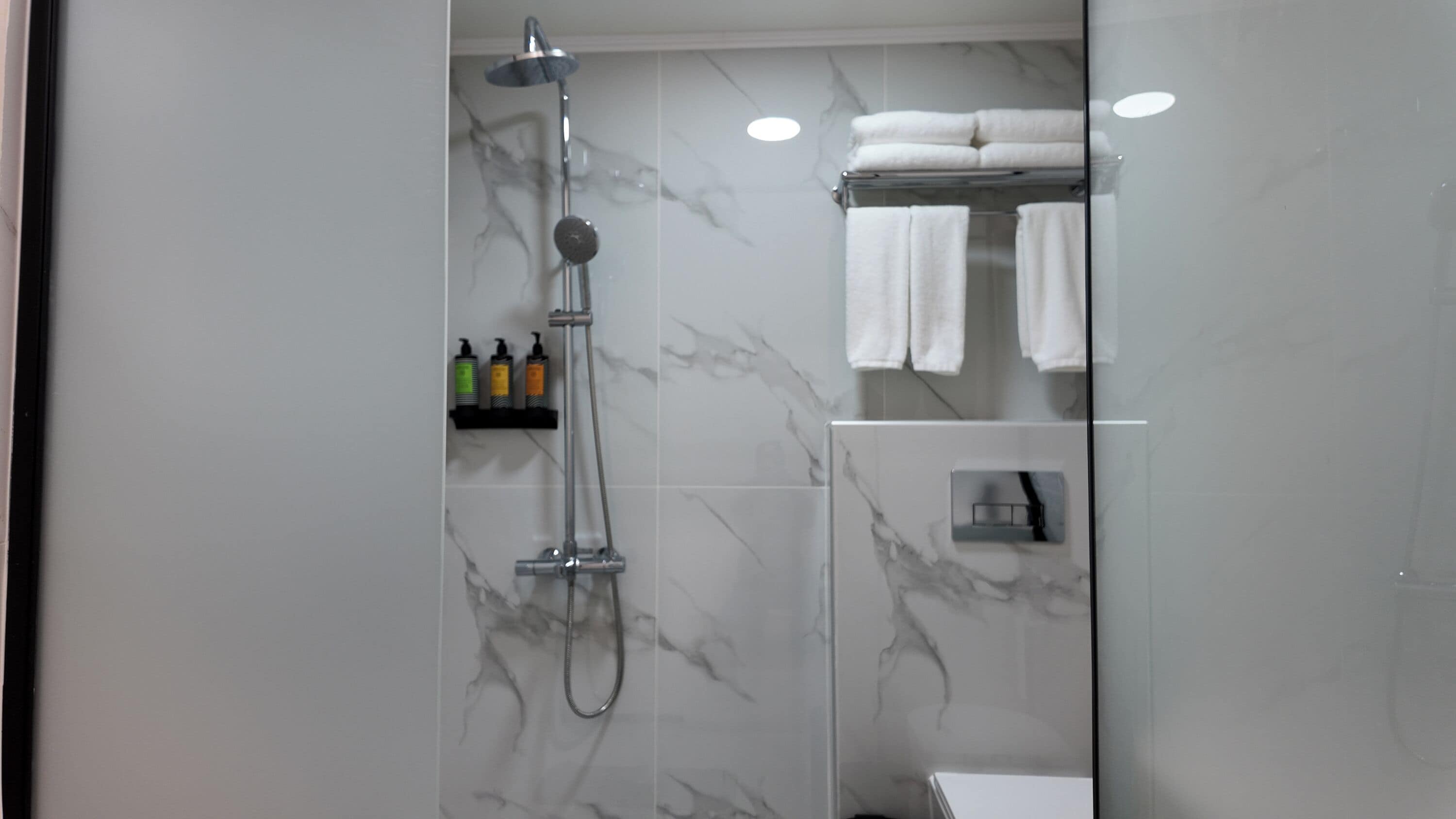 A modern bathroom with a glass shower enclosure, marble walls, and a shelf with toiletries.