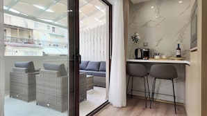Un appartement moderne doté d'un balcon, de meubles en osier, d'un canapé, d'un coin cuisine avec une machine à café et d'une bouteille de vin.