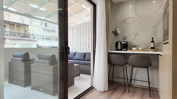 A modern apartment with a balcony, wicker furniture, a sofa, a kitchen area with a coffee machine, and a wine bottle.