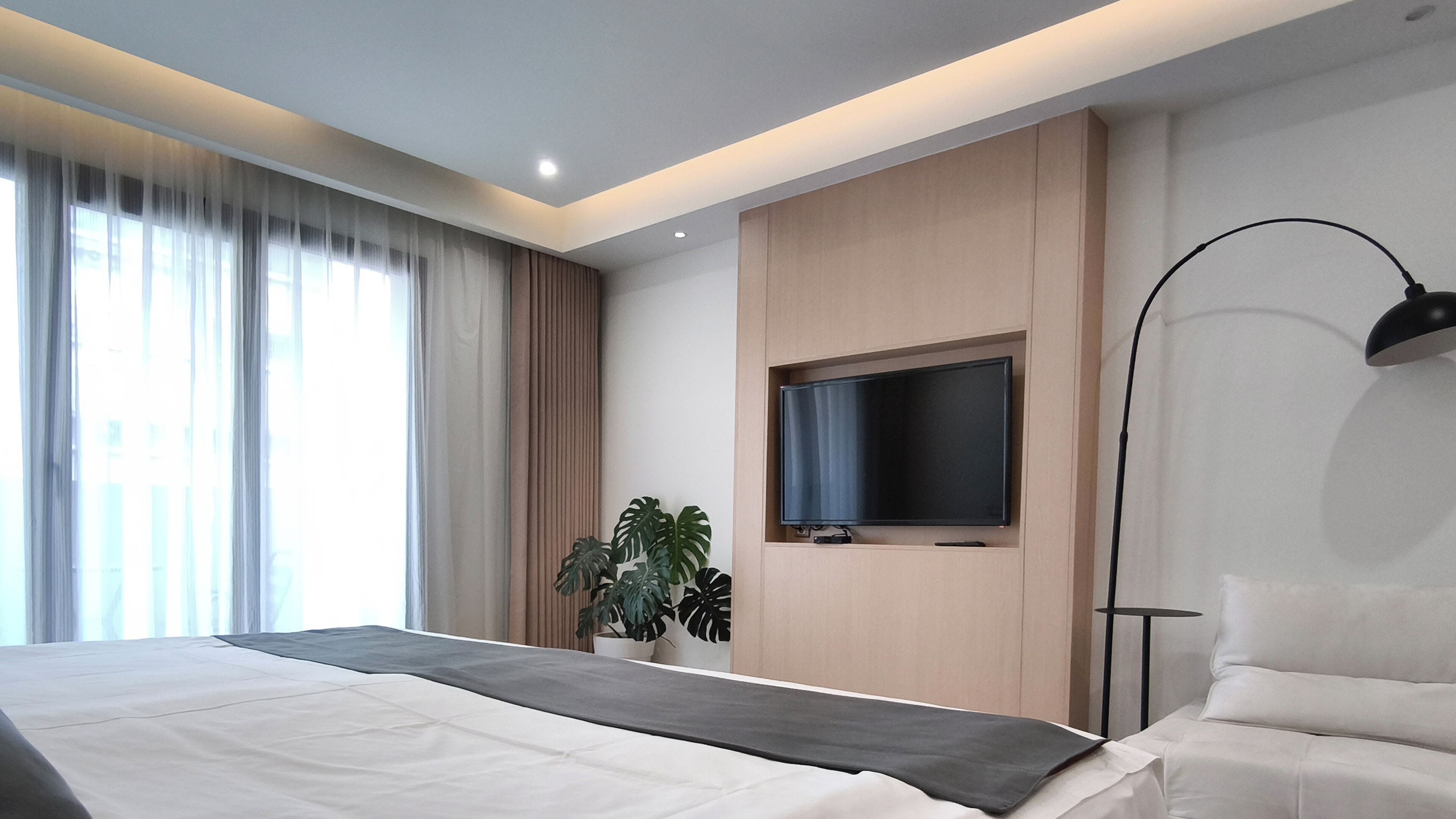 A modern hotel room with a large bed, a flat-screen TV mounted on the wall, a floor lamp, and a potted plant.