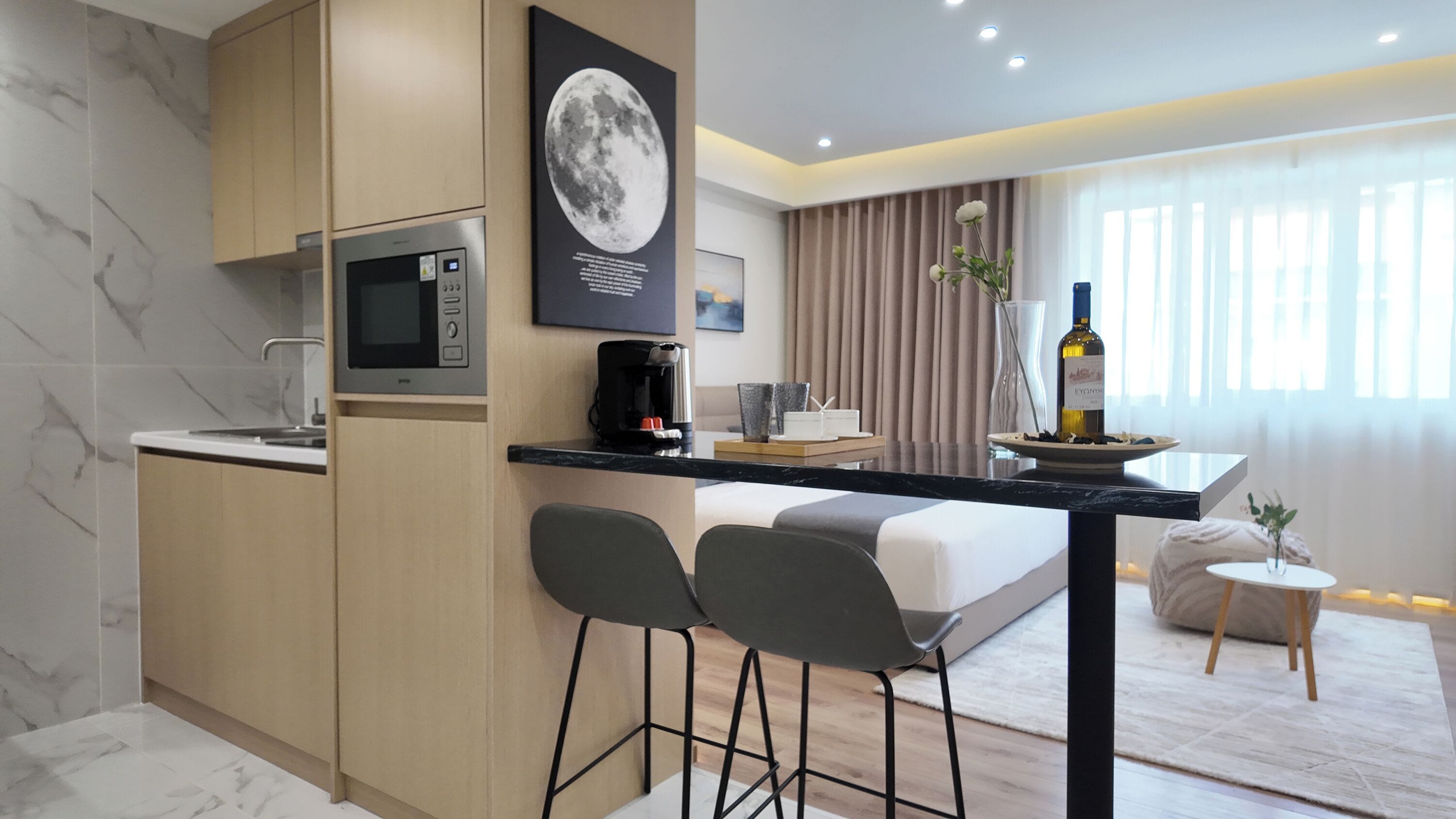A modern hotel room with a kitchenette, a bed, a dining area, and a large window with curtains.