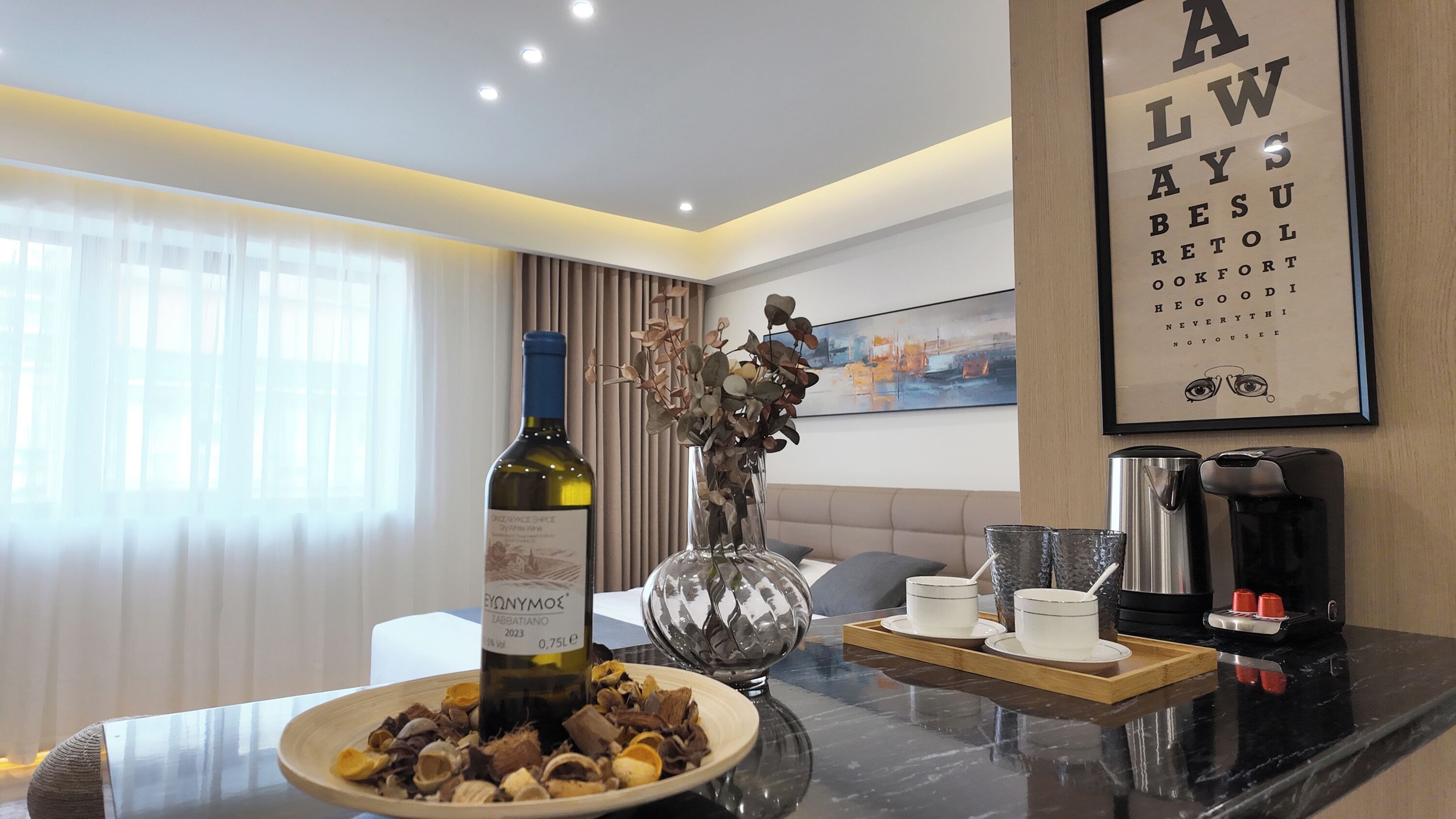 A modern hotel room with a dining area featuring a bottle of wine, a decorative vase, and a coffee machine.