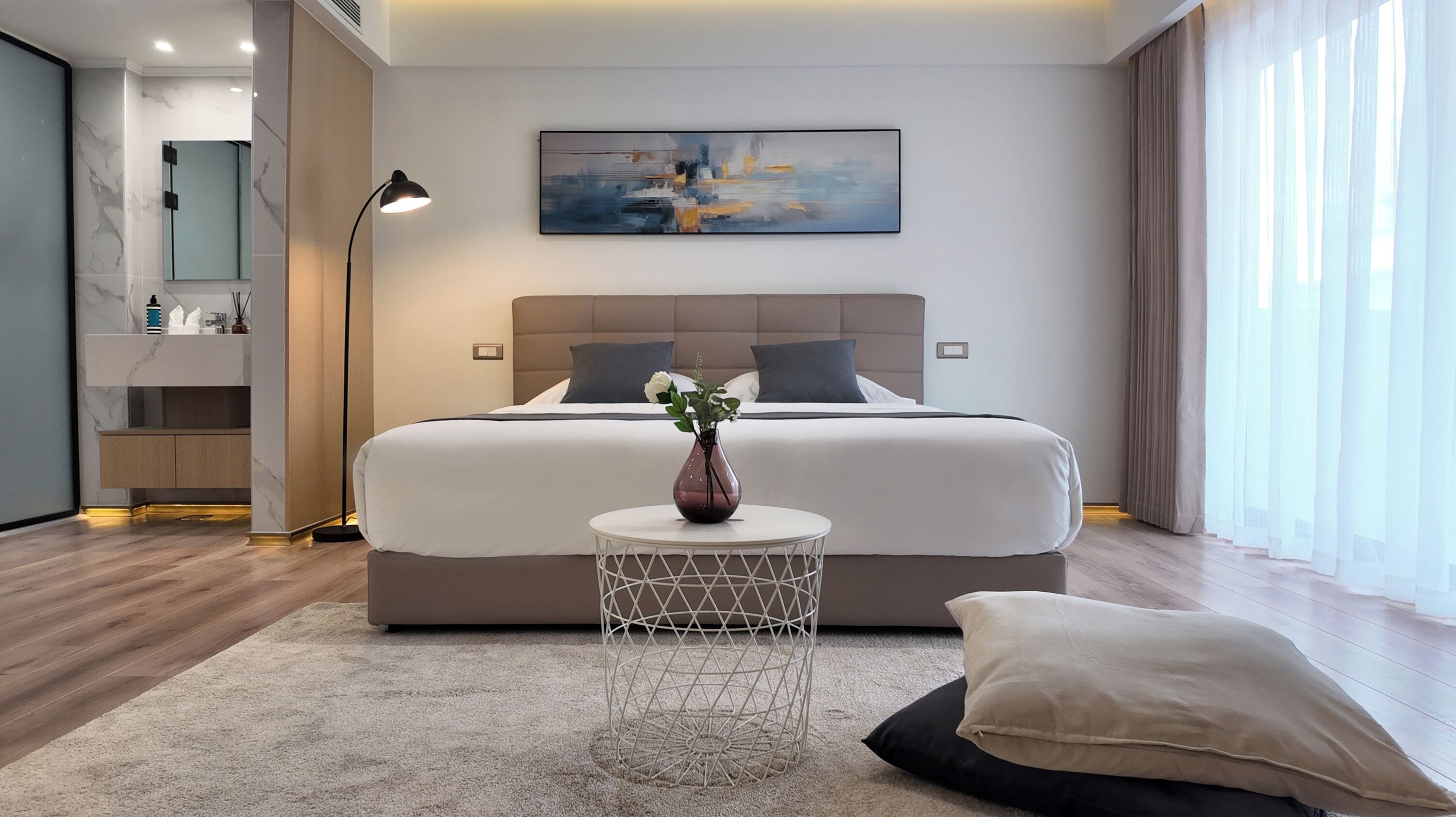 Una habitación de hotel moderna con una cama grande, una mesita de noche con un florero, una mesita redonda y un ventanal con cortinas.