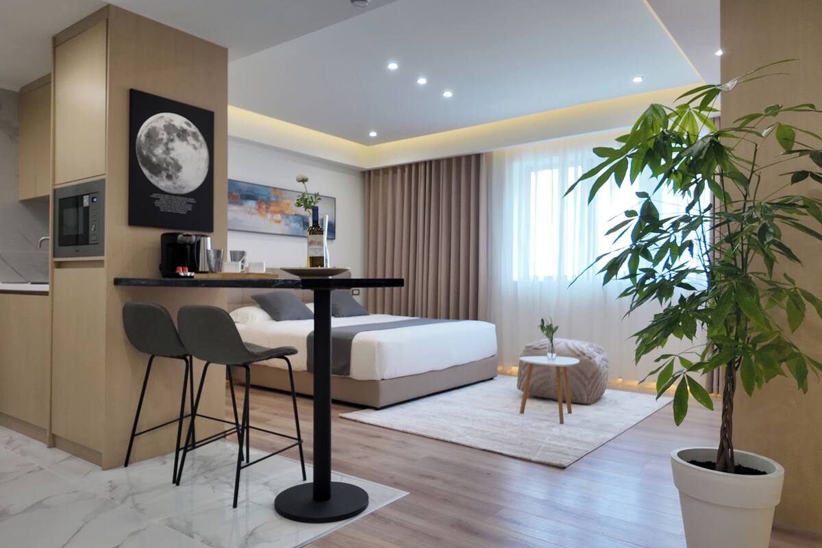 Una habitación de hotel moderna con cama, kitchenette pequeña, zona de comedor y una planta grande en maceta.