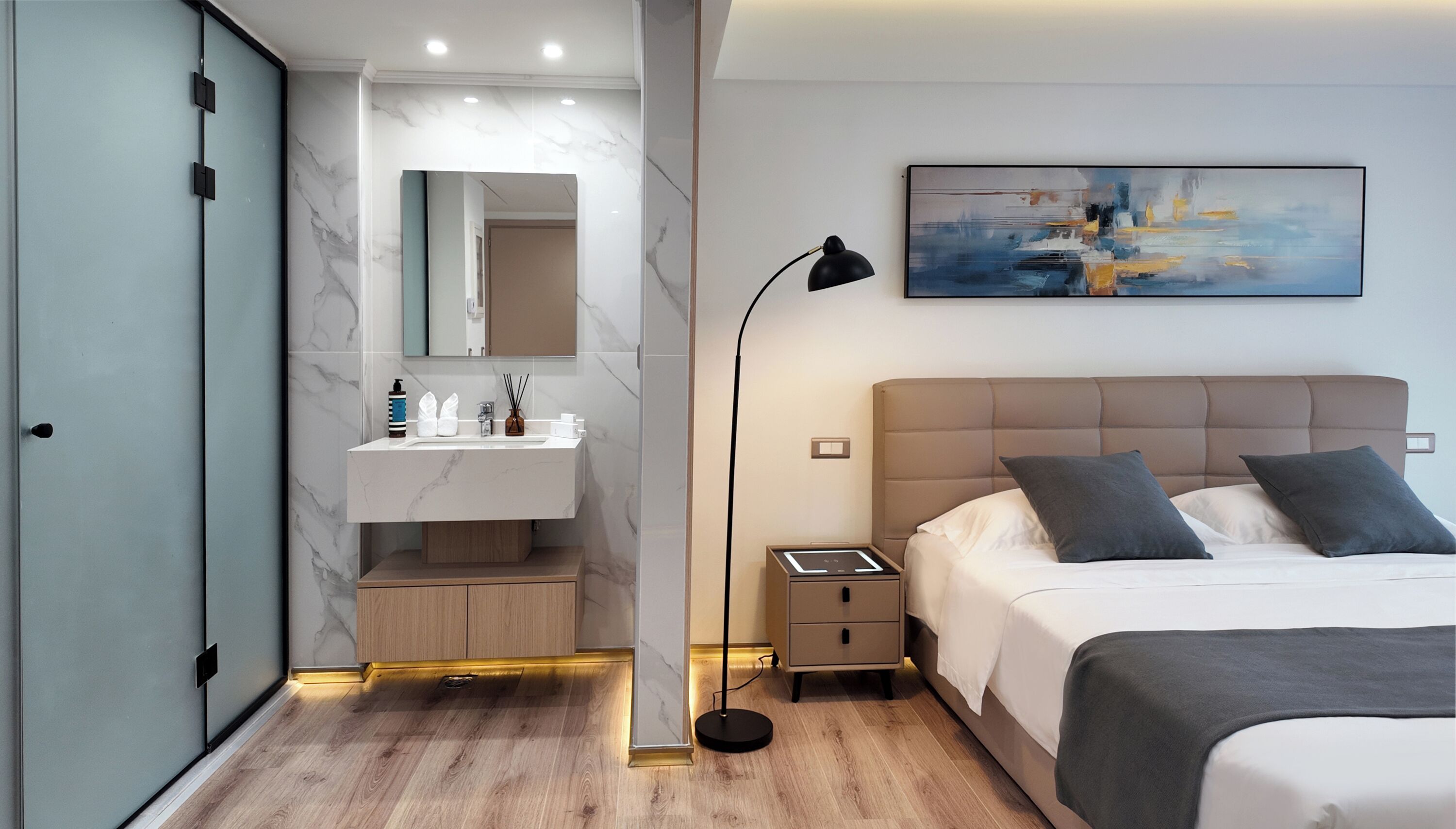 A modern bedroom with a bed, bedside table, and a nightstand. There is a bathroom with a sink, mirror, and storage cabinets.