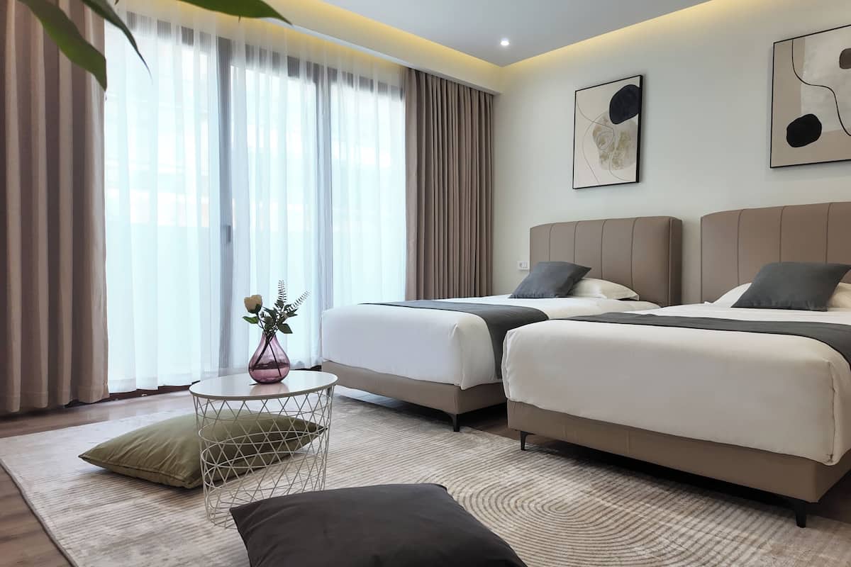 Una habitación de hotel moderna con dos camas, una mesita redonda con un florero y vistas a la ciudad a través de amplias ventanas.