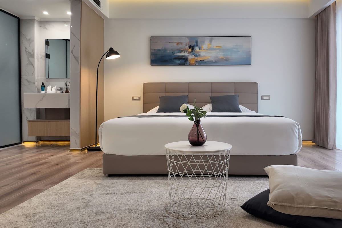 Habitación de hotel moderna con una cama grande, una mesita redonda con un florero, una mesita de noche, un espejo y un cuadro en la pared.