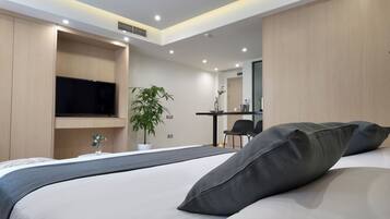 A modern hotel room with a large bed, a flat-screen TV mounted on the wall, a desk with a chair, and a potted plant.