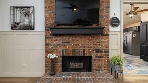 A living room with a brick fireplace, a mounted flat-screen TV, and a ceiling fan. - 
Pool, Game Room + Office - Near the Frisco Star (Frisco)