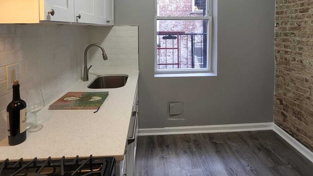Una cocina con armarios blancos, un fregadero de acero inoxidable, una ventana con vista a un edificio de ladrillo y una estufa a gas.