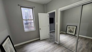 Una habitación con paredes grises, una puerta blanca, una ventana con vista a edificios y un cuadro en la pared.