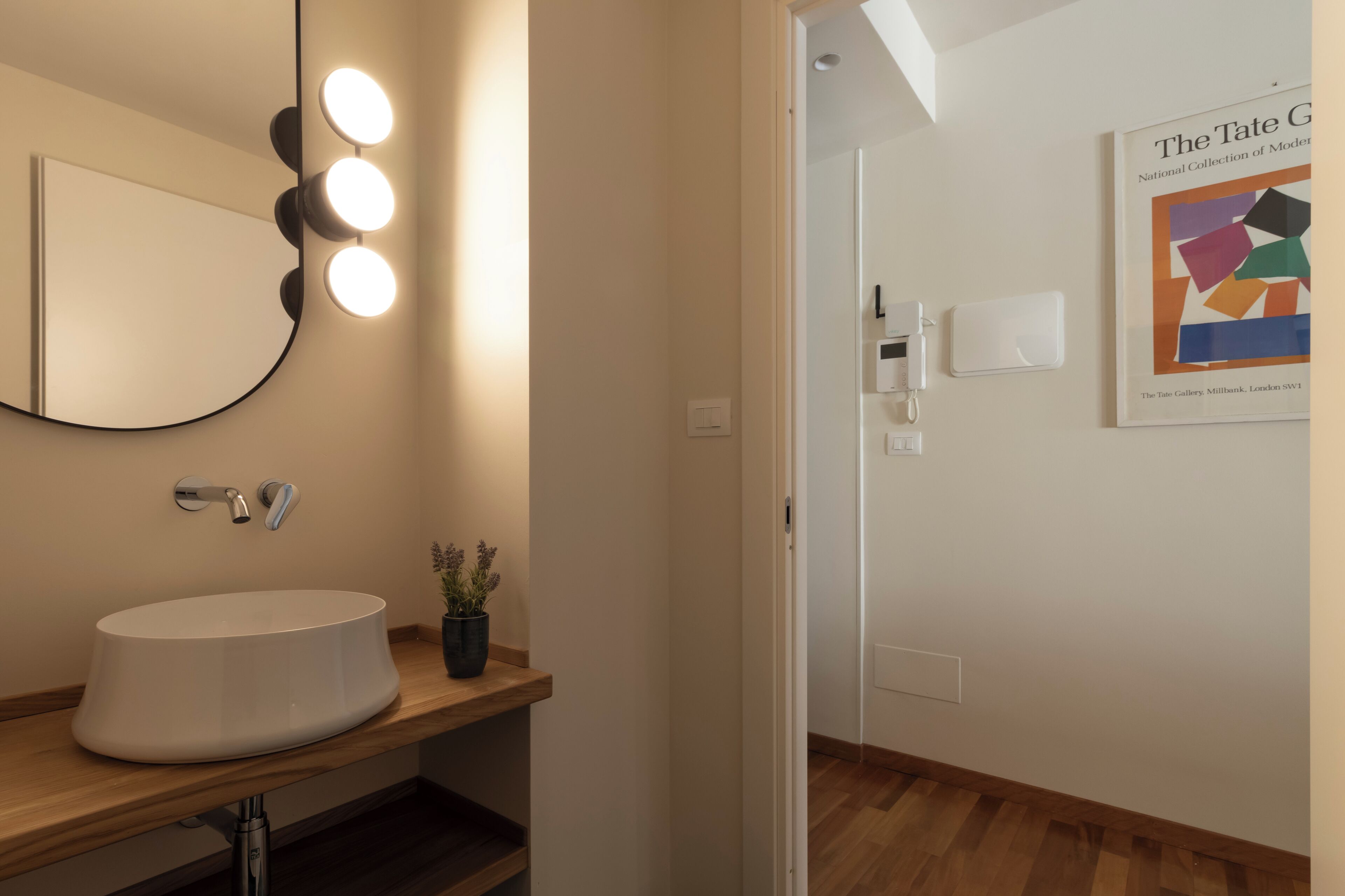 Un baño con un lavabo redondo, un espejo con iluminación, una planta en maceta y un cartel en la pared.