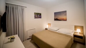 Hotellihuone, jossa on sänky, yöpöytävalaisimet, televisio, patteri, ikkuna verhoilla sekä kaksi seinätaideteosta.