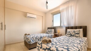 A hotel room with two beds, a window with sheer curtains, an air conditioning unit, and a bedside table with a lamp. - Narcissos Nissi Beach Apartment D12 (Ayia Napa)