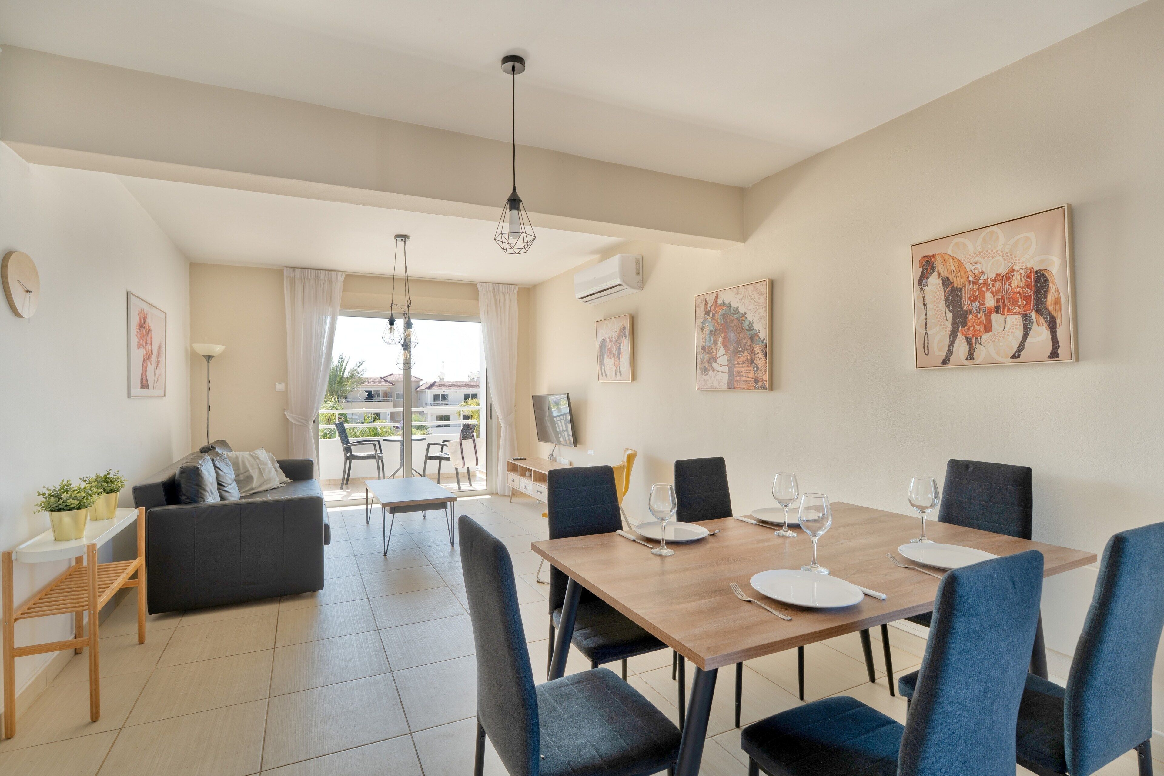 Un espace de salle à manger moderne avec une table en bois dressée pour quatre personnes, entourée de chaises bleues. On aperçoit un canapé et une petite table avec une lampe et une plante au premier plan.