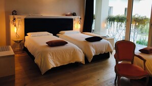 Ein Hotelzimmer mit zwei Betten, Holzboden, einem großen Fenster mit Blick auf ein Gebäude und Grünflächen, und einem roten Sessel.