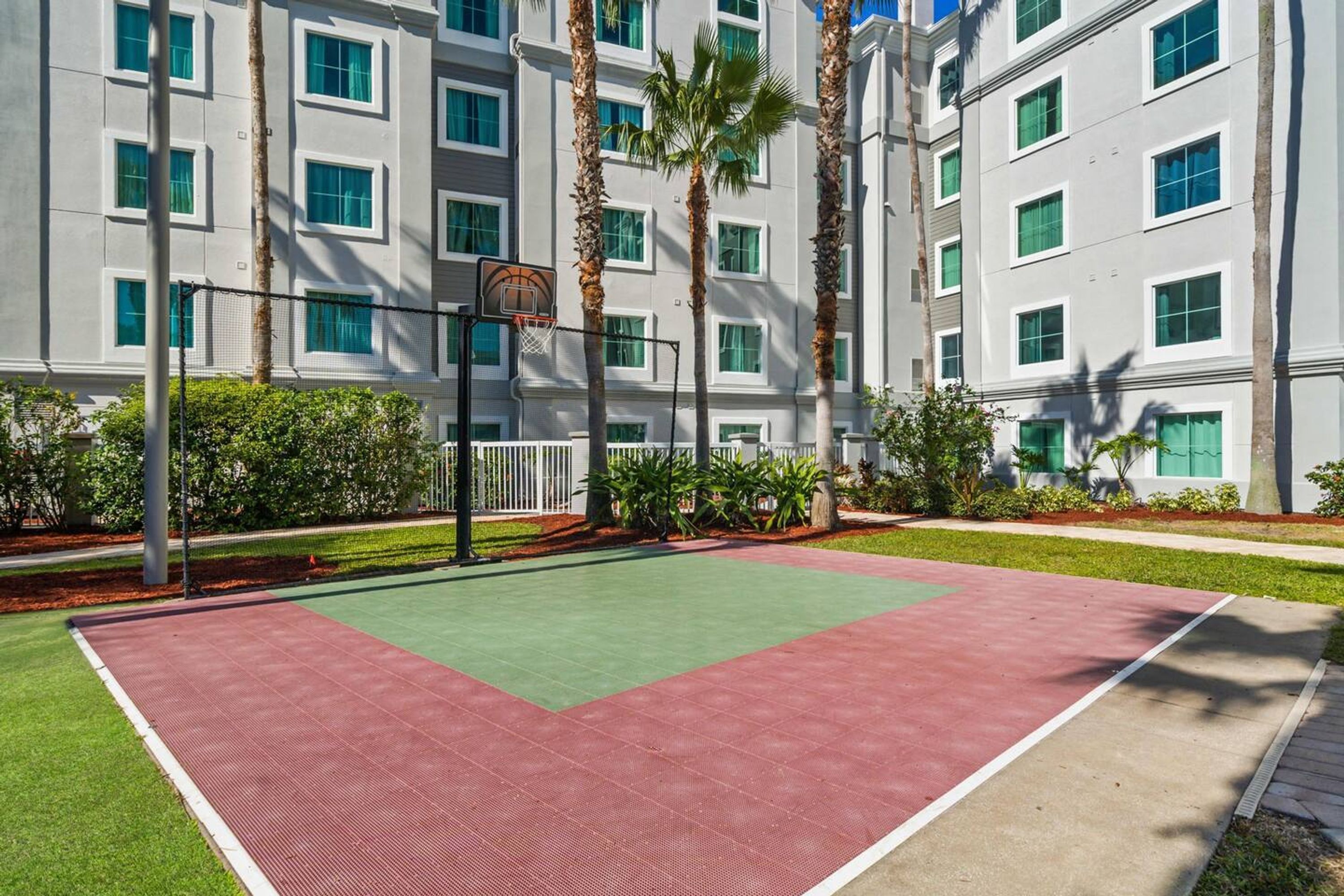 Un terrain de basket en plein air, doté d’une surface verte et rouge, entouré d’un bâtiment avec de nombreuses fenêtres et de palmiers.