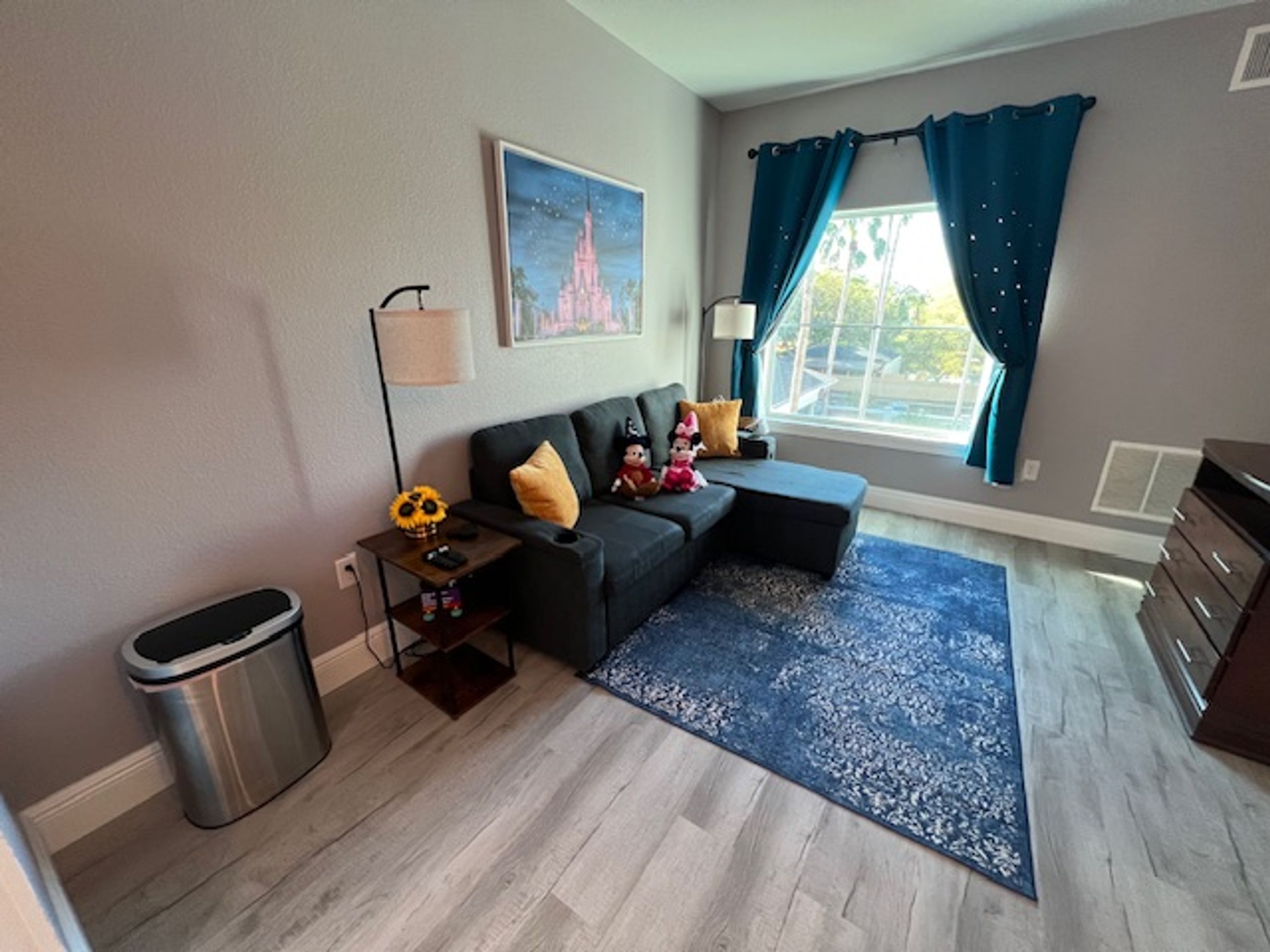 A modern living room with a sofa, a small table with a lamp, a trash bin, and a large window with curtains.