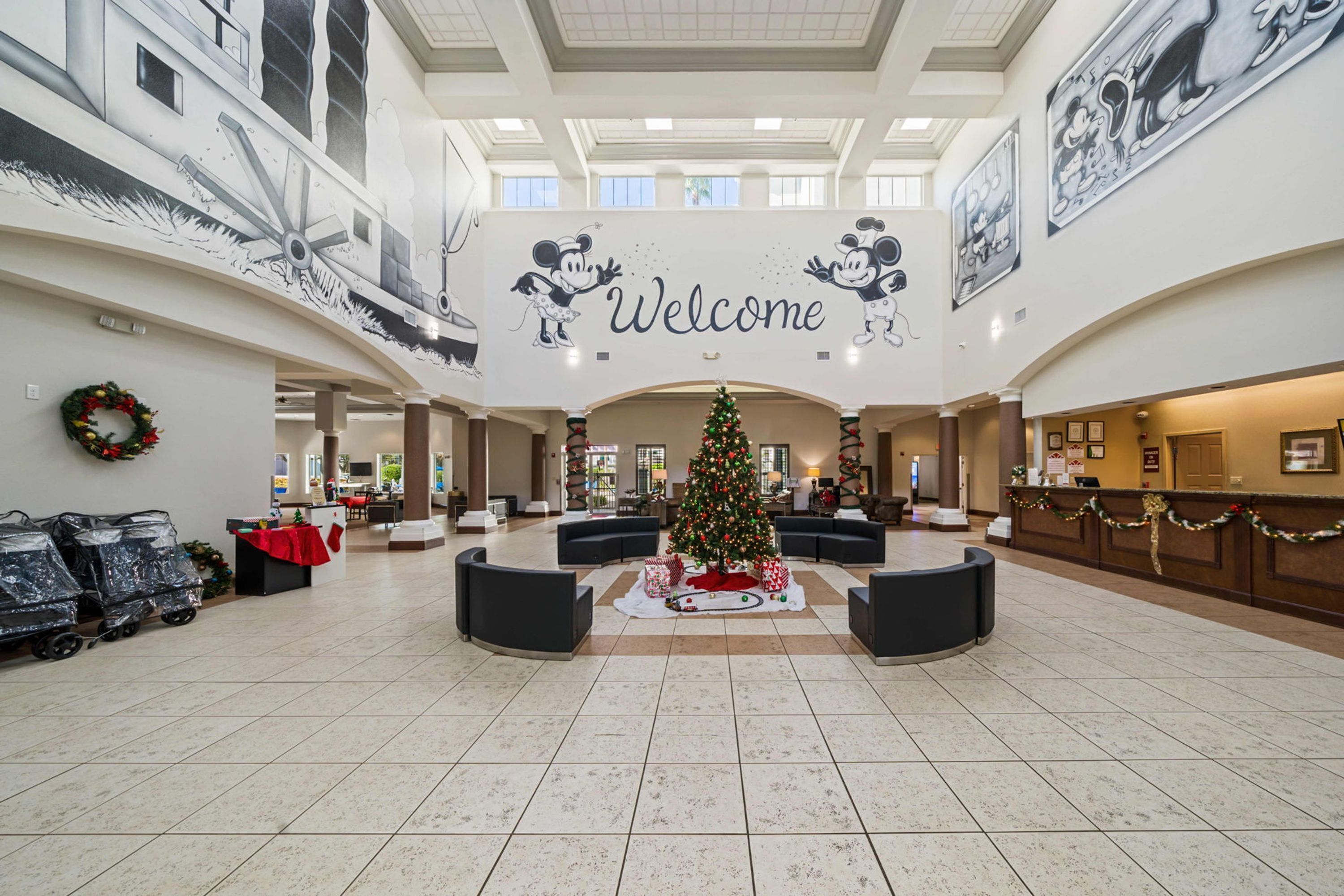 A spacious lobby with a central Christmas tree, a 'Welcome' sign, and Mickey Mouse murals on the walls.
