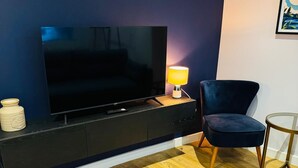 Una sala de estar moderna con un televisor de pantalla plana sobre una consola de madera oscura, un sillón azul y una mesita redonda con una lámpara.