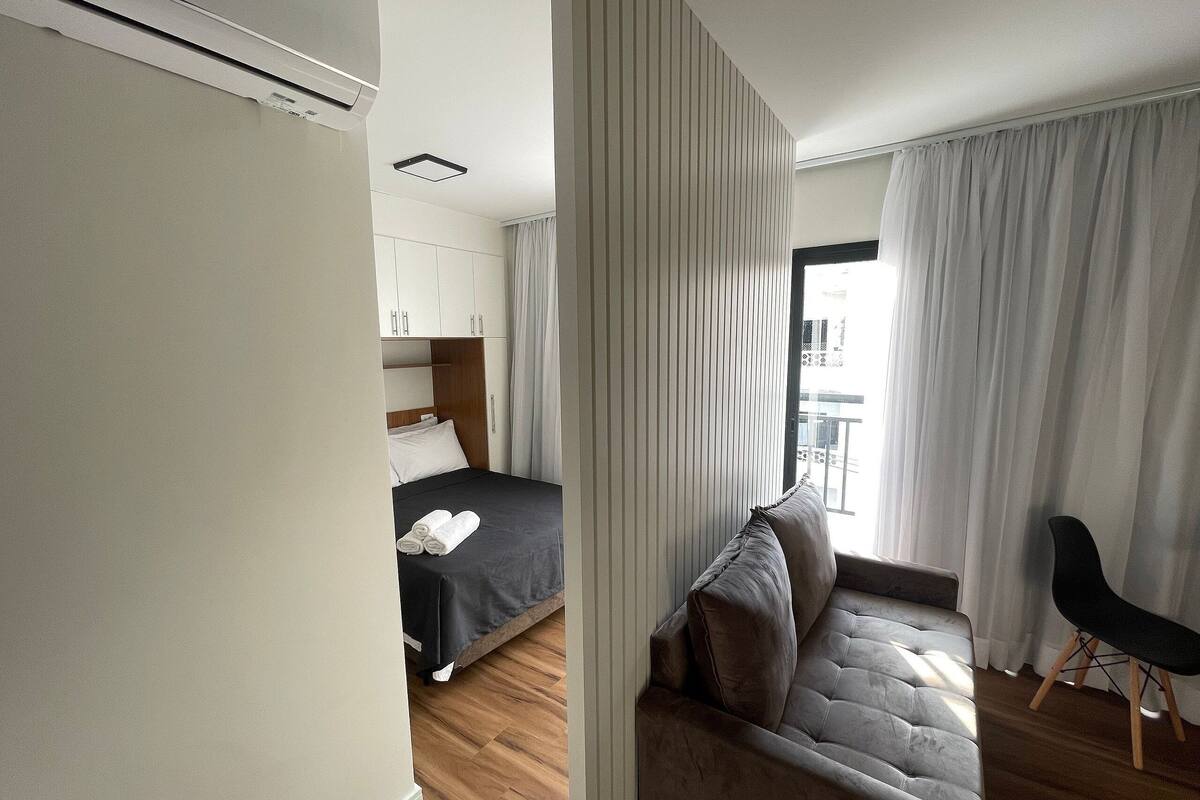 Una habitació d'hotel moderna amb un llit, un sofà i una cadira. Hi ha una finestra amb cortines, un ventilador de sostre i aire condicionat fixat a la paret.