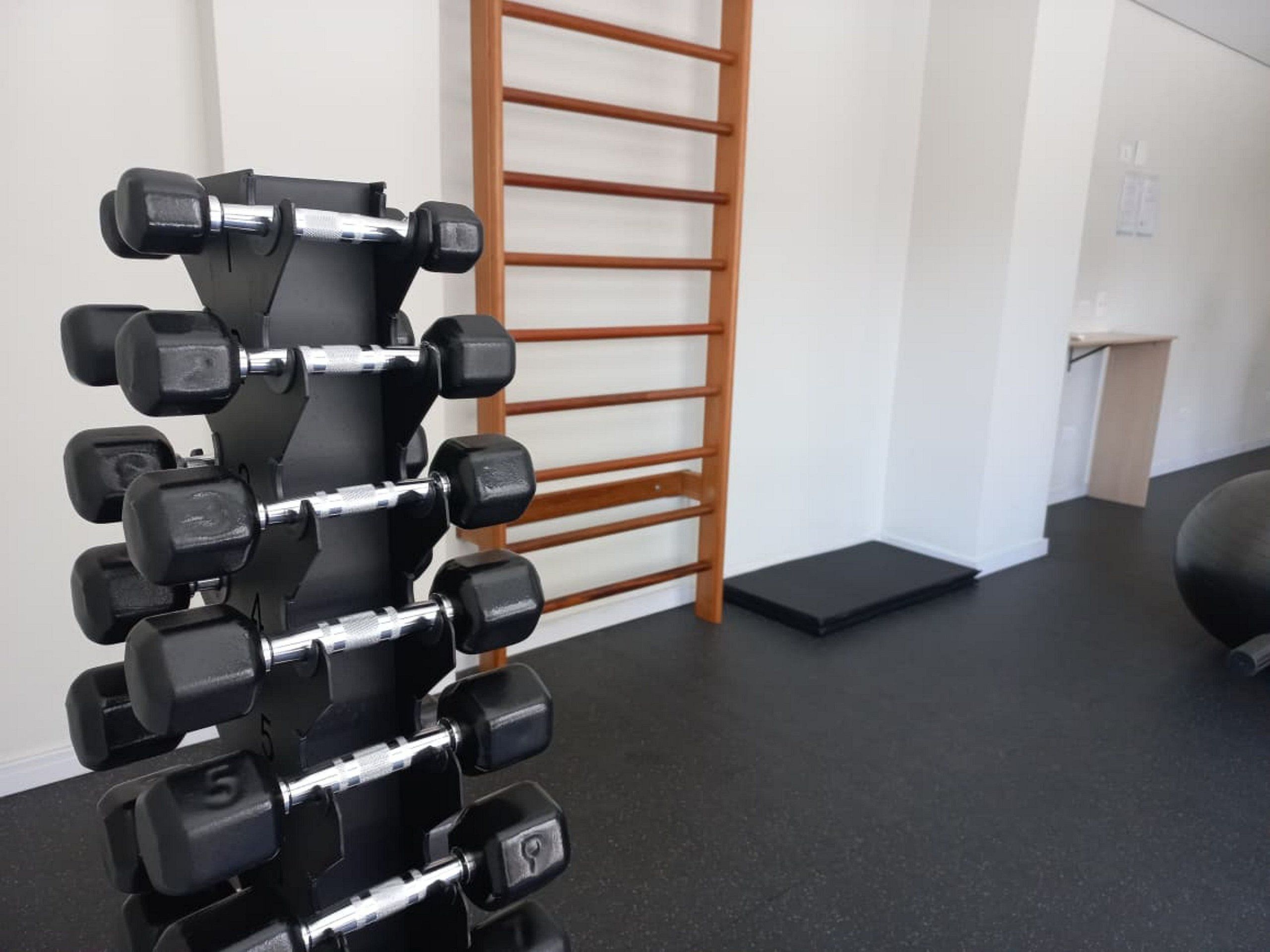 Uma academia com um suporte de halteres fixado na parede, uma escada de madeira e um tapete preto no chão.