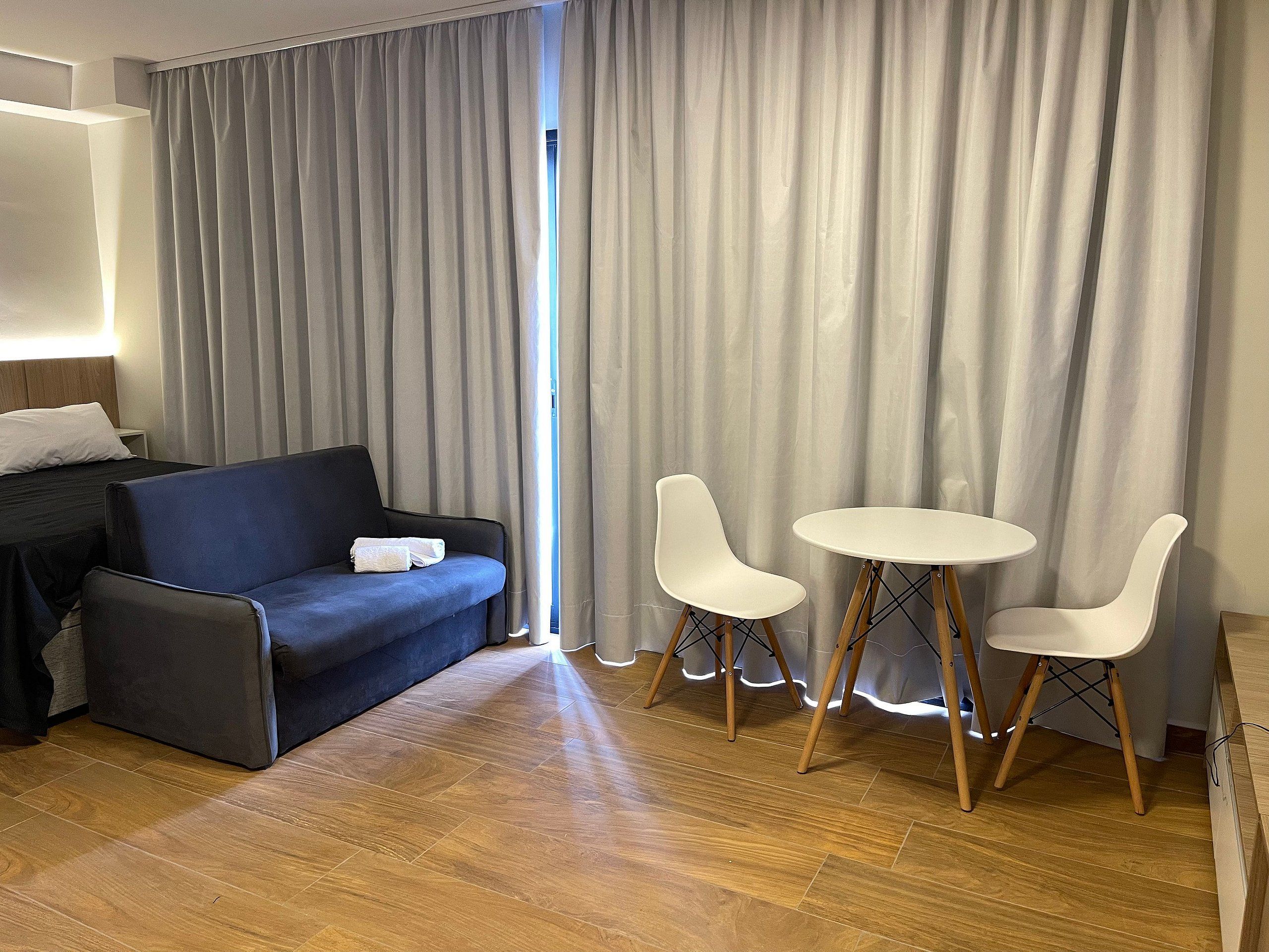 Een moderne hotelkamer met een donkerblauwe bank, twee witte stoelen, een klein rond tafeltje en een groot raam met grijze gordijnen.