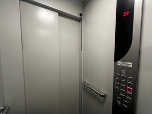 L’intérieur d’un ascenseur est doté d’une porte métallique, d’un panneau de commande avec un bouton rouge d’urgence, et d’un panneau indiquant une capacité maximale de 600 kg et de 8 personnes.