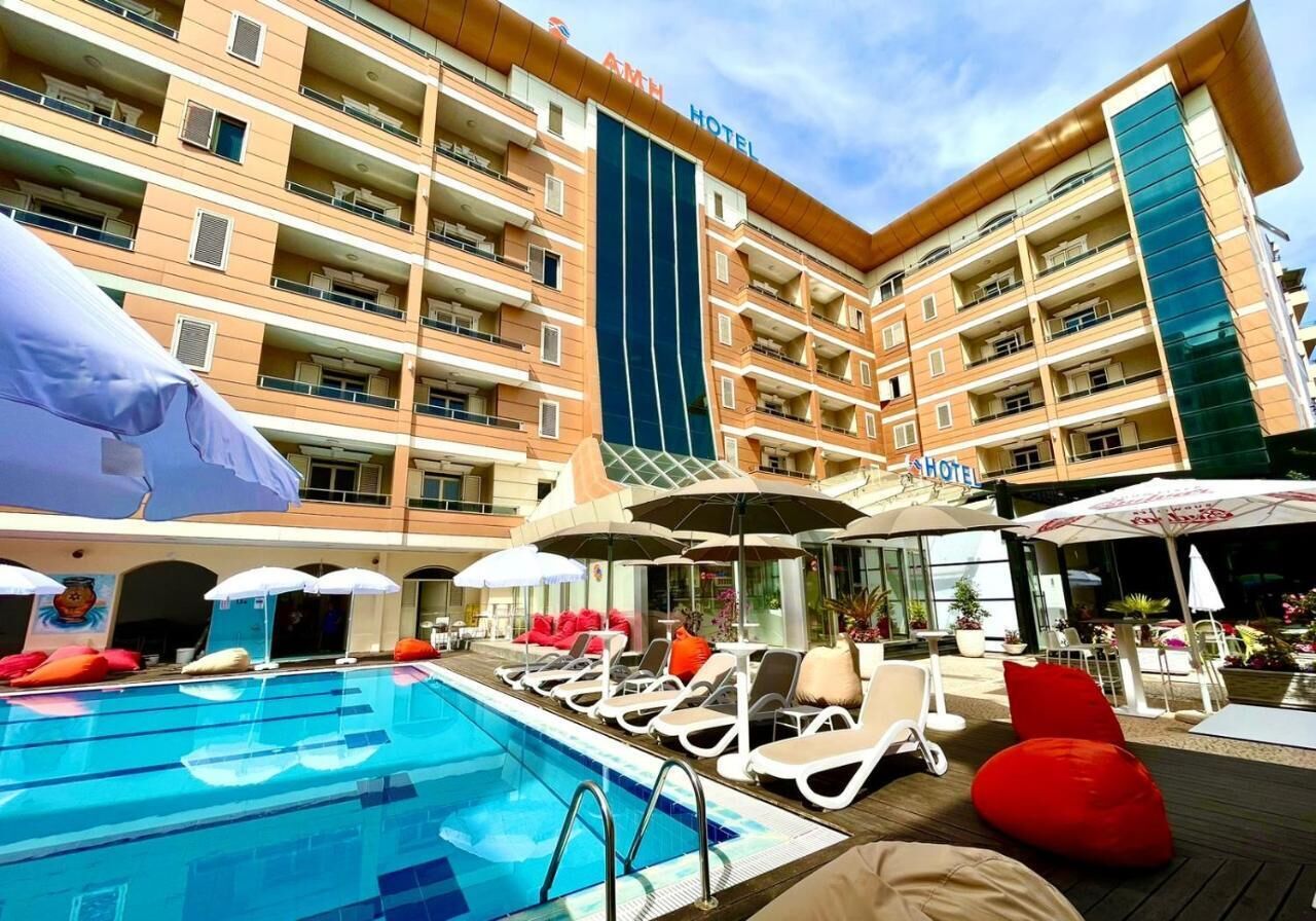 Zona de piscina hotelera con tumbonas y sombrillas, rodeada por un edificio de varios pisos con balcones.